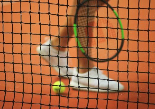 A tennis racket, ball, and player's shoes on a clay court are seen through a tennis net, which is in focus in the foreground.