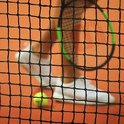 A tennis racket, ball, and player's shoes on a clay court are seen through a tennis net, which is in focus in the foreground.