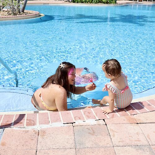 A woman and a child enjoy time by a pool, with the woman in the water and the child sitting on the edge with a toy.