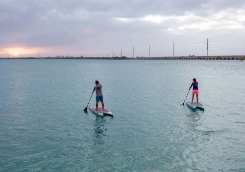 Two people are stand-up paddleboarding on a large body of water at sunset with a long bridge and some trees in the distance.