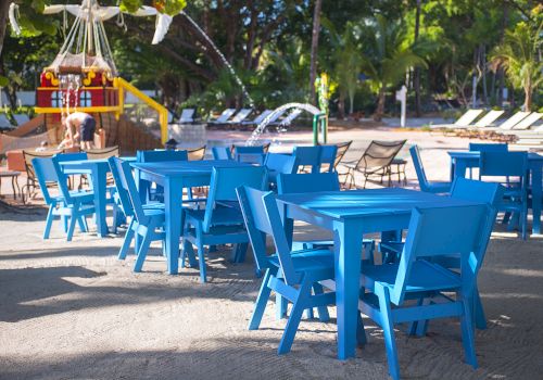 The image shows blue tables and chairs arranged outdoors, with trees, lounge chairs, and a playground structure in the background.