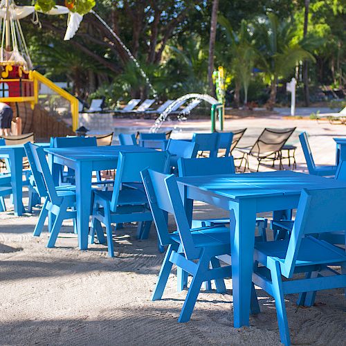 The image shows blue tables and chairs arranged outdoors, with trees, lounge chairs, and a playground structure in the background.