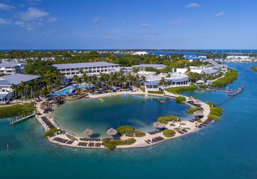 An aerial view of a tropical resort featuring a lagoon, surrounded by multiple buildings, a swimming pool, palm trees, and clear blue water.