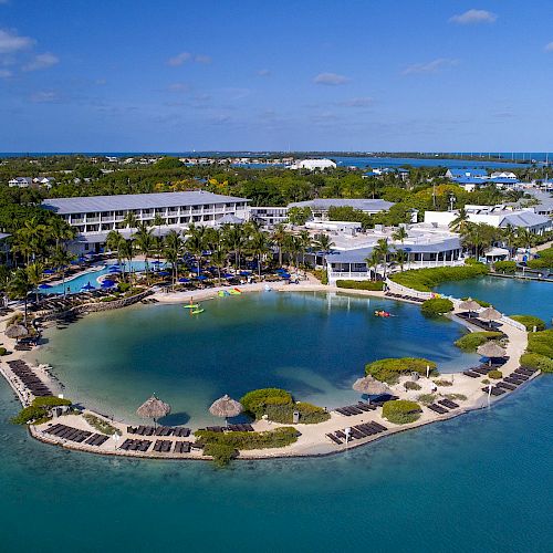 An aerial view of a tropical resort featuring a lagoon, surrounded by multiple buildings, a swimming pool, palm trees, and clear blue water.