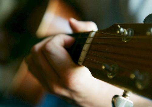 A person is playing an acoustic guitar, with a close-up view of their hand on the fretboard. The image focuses on the guitar strings and fingers.