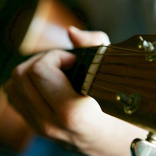 A person is playing an acoustic guitar, with a close-up view of their hand on the fretboard. The image focuses on the guitar strings and fingers.