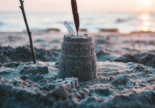 A small sandcastle topped with a feather and a stick nearby, set on a beach during sunset with waves in the background ending the sentence.