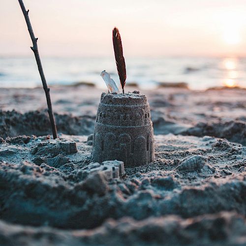 A small sandcastle topped with a feather and a stick nearby, set on a beach during sunset with waves in the background ending the sentence.