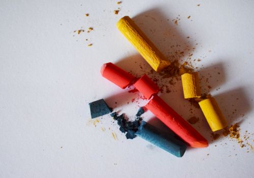 The image shows broken pieces of colorful chalk in red, yellow, and blue shades scattered on a white surface, with some chalk dust around them.