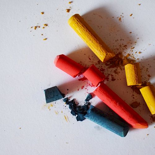 The image shows broken pieces of colorful chalk in red, yellow, and blue shades scattered on a white surface, with some chalk dust around them.