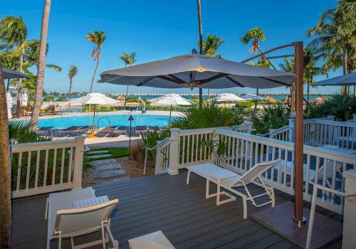 A tropical resort scene with lounge chairs, umbrellas, palm trees, and a pool seen from a deck. The atmosphere is inviting and relaxing.