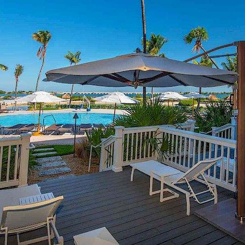 A tropical resort scene with lounge chairs, umbrellas, palm trees, and a pool seen from a deck. The atmosphere is inviting and relaxing.