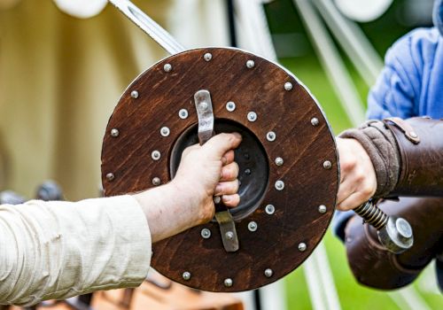 Two people, one in medieval-style clothing, are holding a wooden shield with metal rims during a historical reenactment or event.