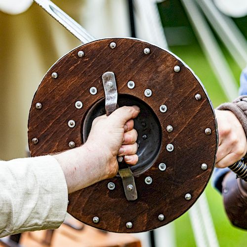 Two people, one in medieval-style clothing, are holding a wooden shield with metal rims during a historical reenactment or event.