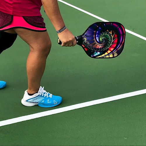 A person in sports attire holding a colorful pickleball paddle on a green court, preparing to play.