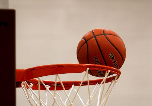 A basketball is seen resting on the rim of a basketball hoop with a net, indicating a close or potential successful shot.