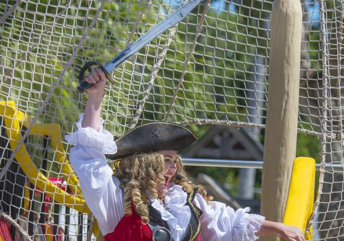 A person dressed as a pirate stands on a colorful play ship, raising a sword triumphantly while wearing traditional pirate attire.