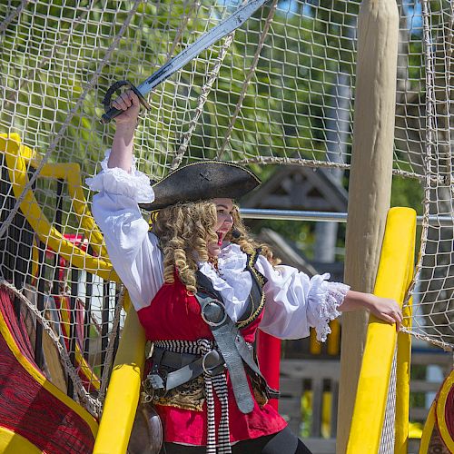 A person dressed as a pirate stands on a colorful play ship, raising a sword triumphantly while wearing traditional pirate attire.