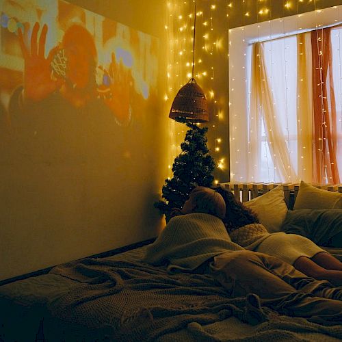 A cozy room with string lights, a projected movie, a bed with people lying in it, a window with curtains, and small Christmas trees.