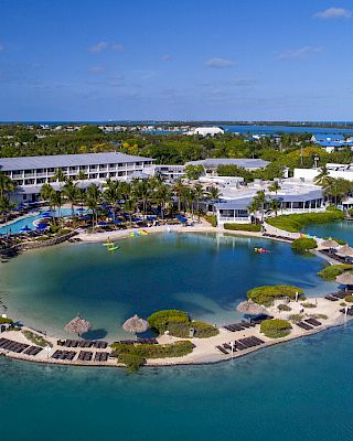 The image shows a resort with buildings, a pool, and a lagoon surrounded by lounge chairs and palm trees, set in a tropical coastal area.