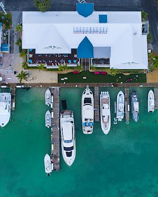 An aerial view of a marina shows several boats docked along a pier beside buildings with white and blue rooftops, adjacent to turquoise water.