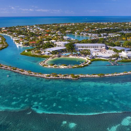 An aerial view of a scenic island surrounded by clear blue waters, featuring greenery, buildings, and a small circular lagoon in the center.