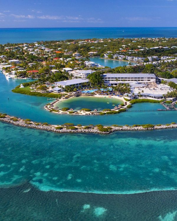 An aerial view of a scenic island surrounded by clear blue waters, featuring greenery, buildings, and a small circular lagoon in the center.