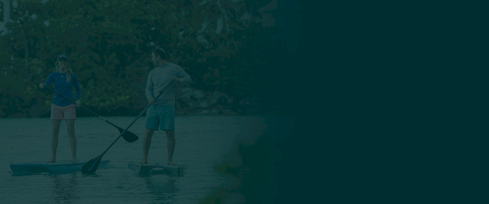 Two people are paddleboarding on a calm body of water with trees in the background. Both are standing on their boards and holding paddles.