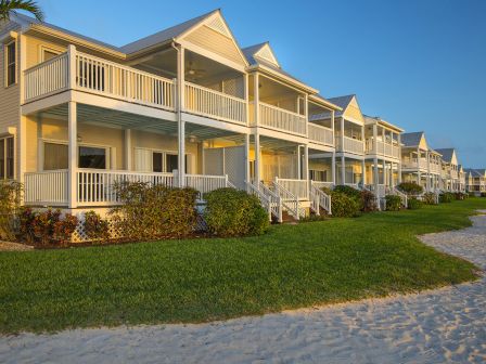 A row of beachfront houses with large porches, lush greenery, and a sandy path leading to the ocean, under a clear sky in the early evening.