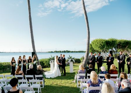 A wedding ceremony is taking place outdoors by the water, with guests seated, the wedding party standing, and the bride and groom at the altar.