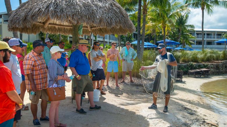 A group of people stands under a thatched roof, watching a man demonstrating how to use a fishing net near a beach with palm trees and buildings.