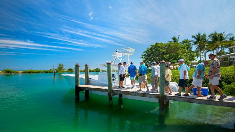 A group of people is standing on a dock by a vibrant turquoise body of water, preparing to board a boat under a clear, sunny sky with scattered clouds.