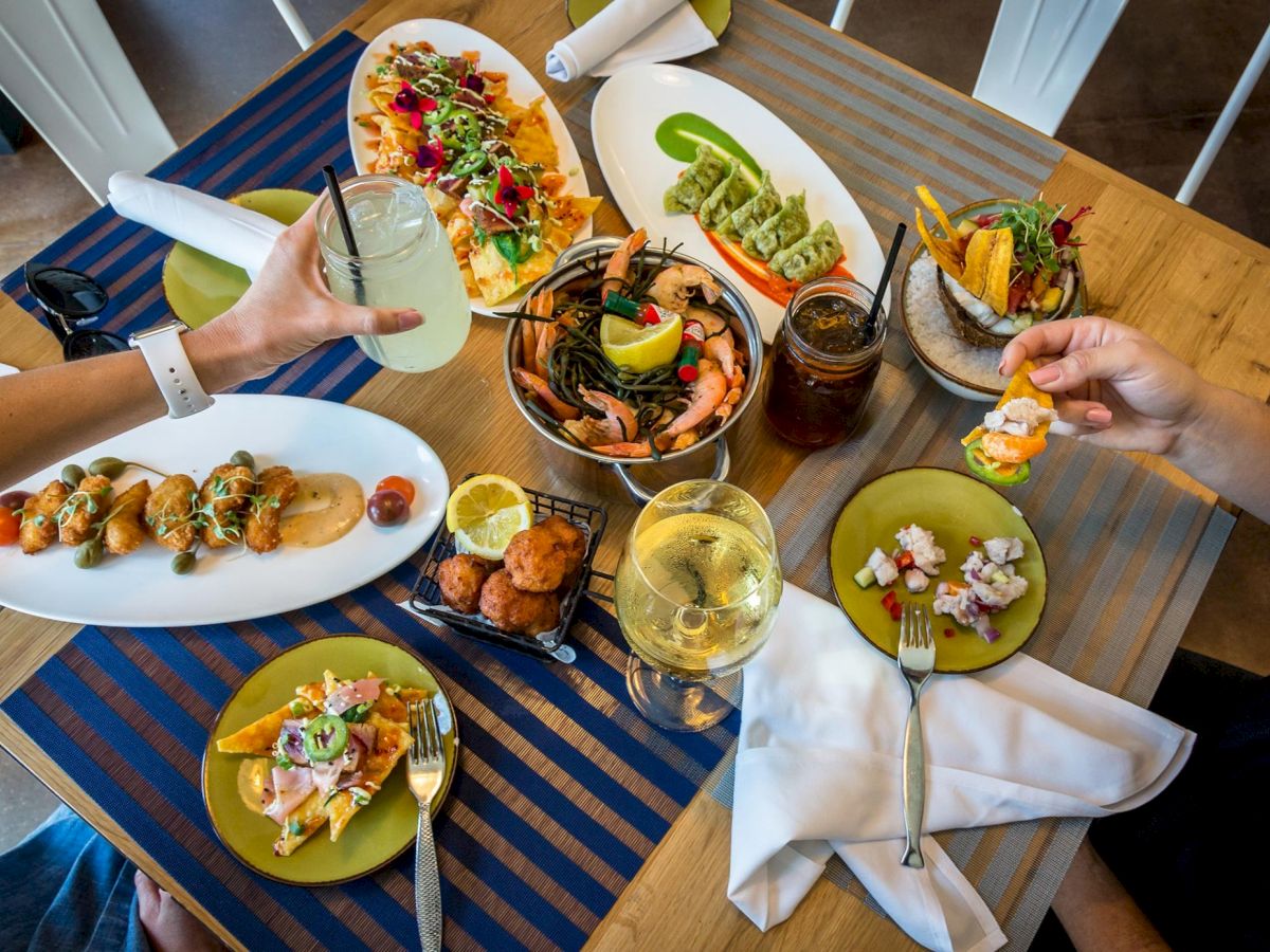 A table filled with various dishes including seafood, tacos, and salads with people enjoying drinks and food in a communal setting.