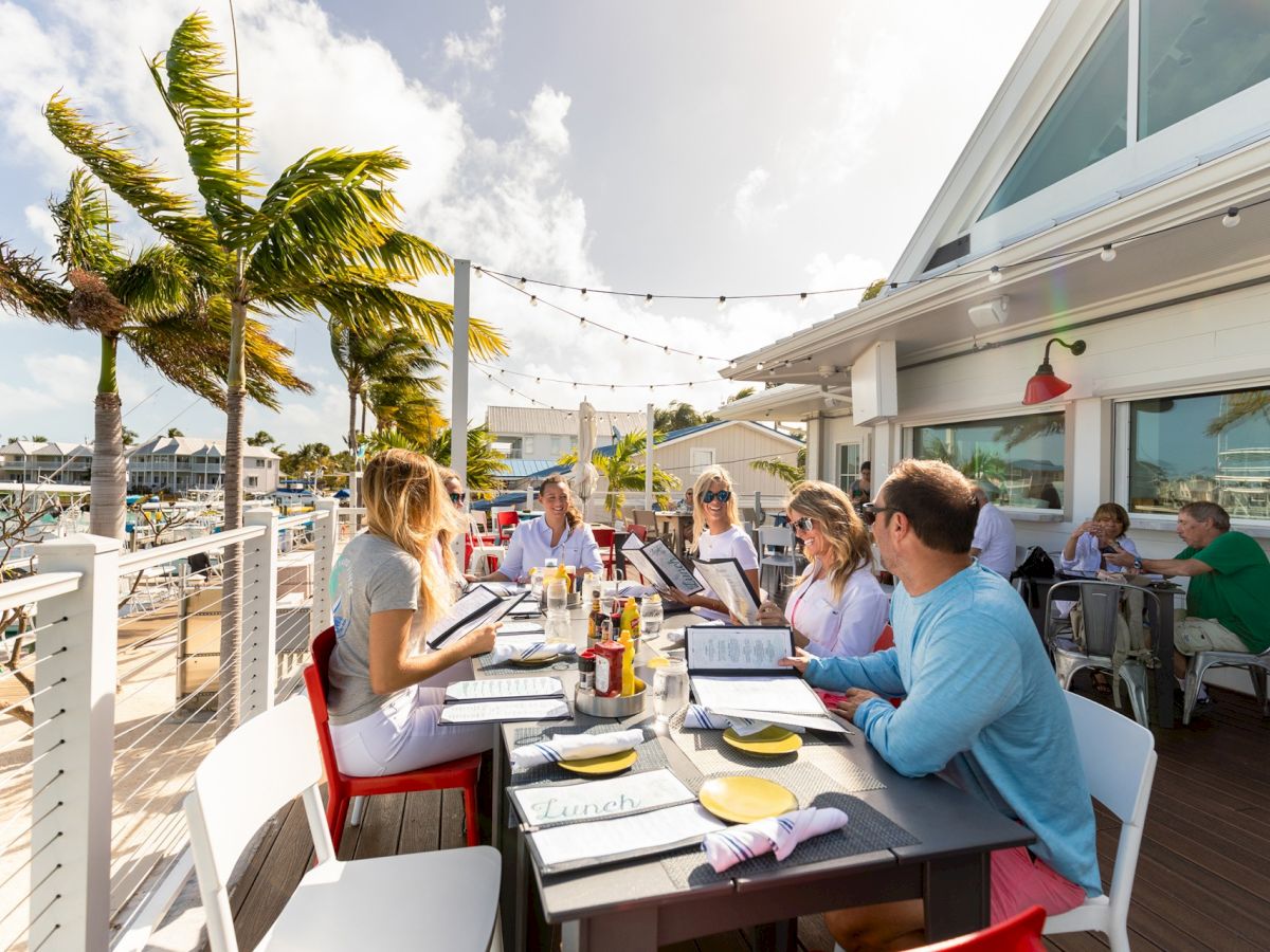 People dining outdoors at a beachside restaurant, with palm trees and string lights in the background, enjoying a sunny day.