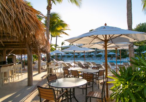 Outdoor dining area with wooden tables, chairs, and parasols next to a bar under thatched roofing. A pool and palm trees are in the background.