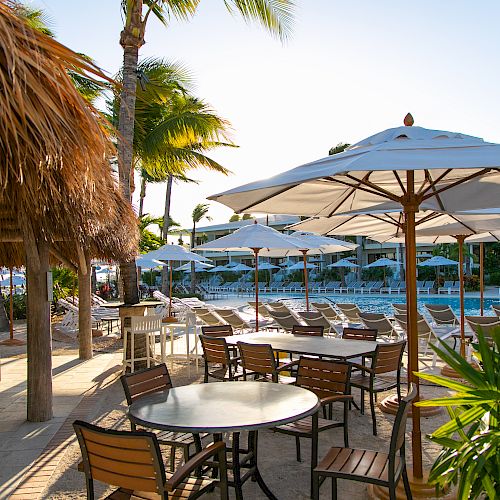 Outdoor dining area with wooden tables, chairs, and parasols next to a bar under thatched roofing. A pool and palm trees are in the background.