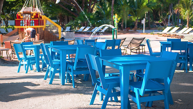 Outdoor setting with blue tables and chairs, a sandy floor, palm trees, a playground, and lounge chairs in the background.