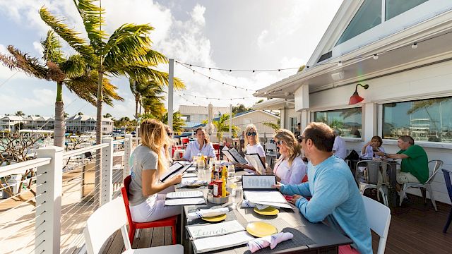 People dining at an outdoor restaurant by the water, with palm trees, string lights, and a bright, sunny atmosphere. Everyone is looking at menus.