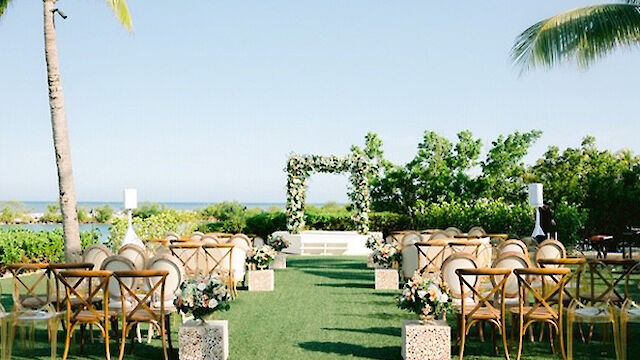 The image shows an outdoor wedding setup with chairs, a floral arch, and palm trees, overlooking a scenic view with greenery and the ocean in the background.