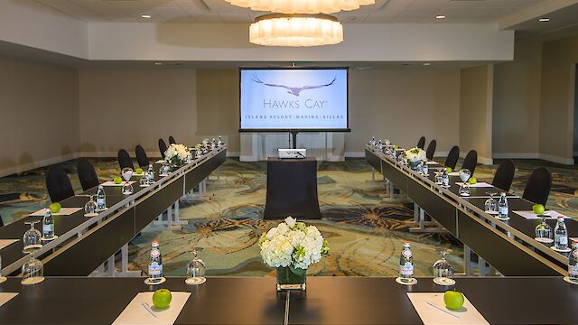 This image shows a conference room set up with U-shaped tables, chairs, water bottles, apples, and flowers, facing a screen displaying 