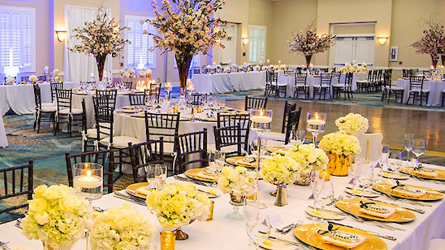 Elegant banquet hall with long tables, yellow-themed decor, floral centerpieces, and candles. Tables set for a formal event with plates and glasses.