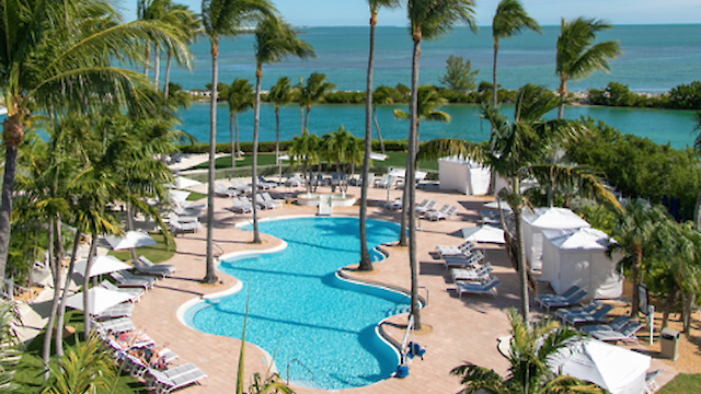 An aerial view of a luxurious resort featuring a pool, palm trees, lounge chairs, cabanas, and a clear ocean in the background, under a blue sky.