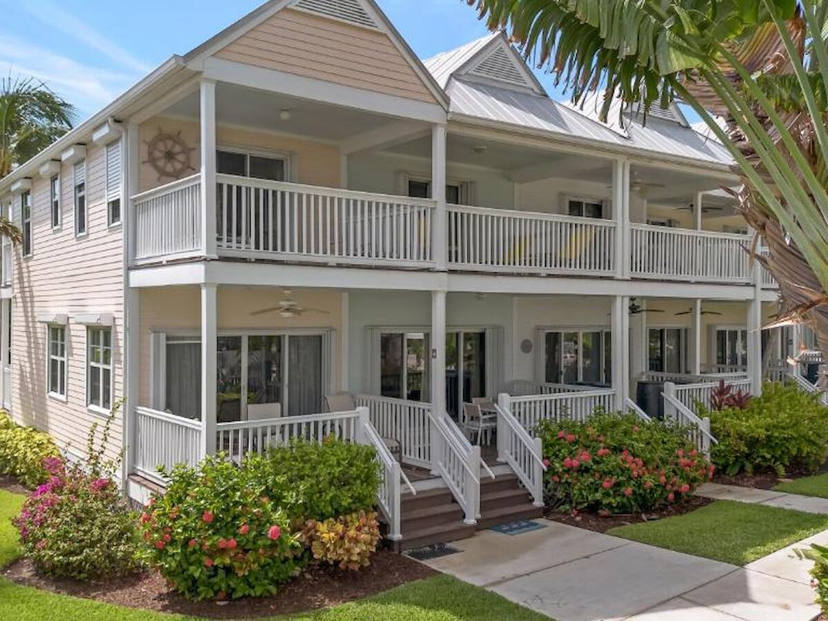 A duplex with large balconies, white railings, and lush landscaping featuring palm trees and flowering shrubs on a sunny day.