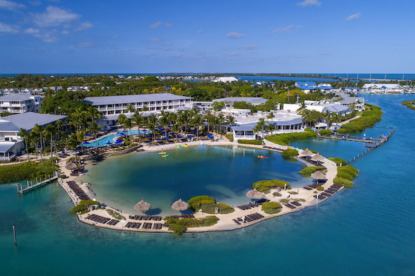 An aerial view of a resort with a large pool, surrounding buildings, palm trees, and waterfront access.