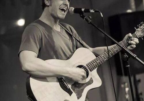 A person is singing into a microphone while playing an acoustic guitar on stage. The image is in black and white.