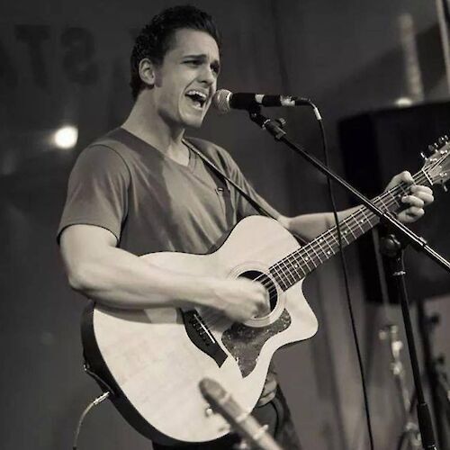 A person is singing into a microphone while playing an acoustic guitar on stage. The image is in black and white.