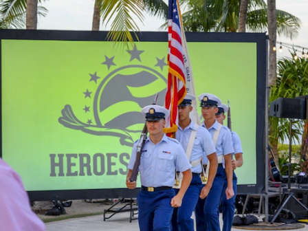 Four uniformed individuals are carrying a flag, marching in front of a large screen displaying the word 