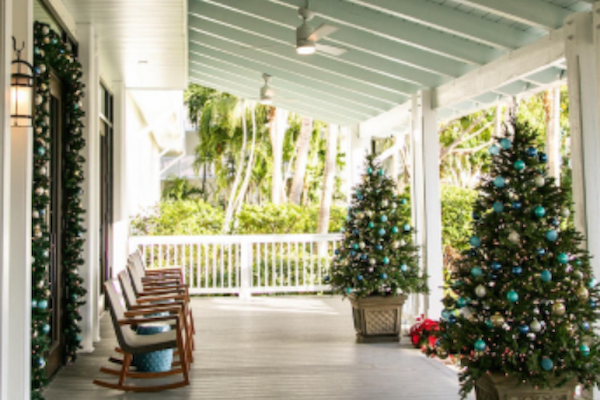 A decorated porch with rocking chairs, Christmas trees adorned with blue and green ornaments, and a lush green background.